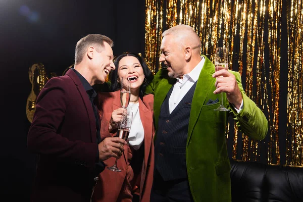 Happy asian woman holding glass of champagne near mature friends during party on black background — Stock Photo