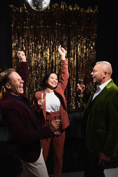 Excited multiethnic friends with champagne showing yes gesture near disco ball and tinsel on black background — Stock Photo