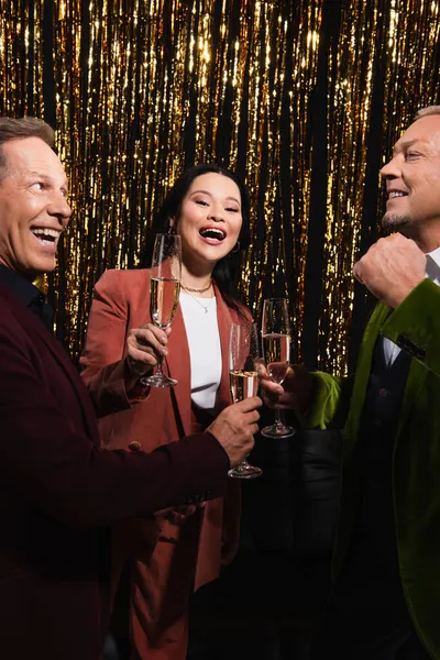 Positive asiatique femme tenant verre de champagne près mature amis et tinsel sur fond noir — Photo de stock