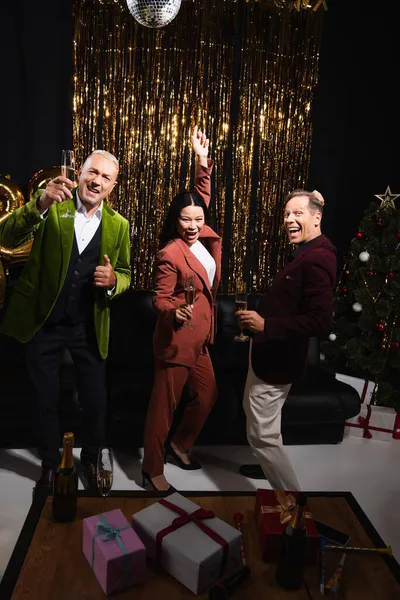 Amigos multiétnicos positivos con champán bailando cerca de regalos y árbol de Navidad sobre fondo negro - foto de stock