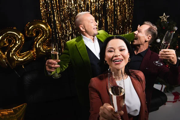 Vue grand angle de la femme asiatique tenant un verre de champagne près des amis et décor festif sur fond noir — Photo de stock