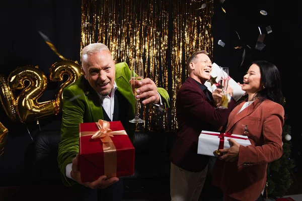 Positive reife Mann mit Champagner und Geschenk in der Nähe interracial Freunde während der Silvesterparty auf schwarzem Hintergrund — Stockfoto