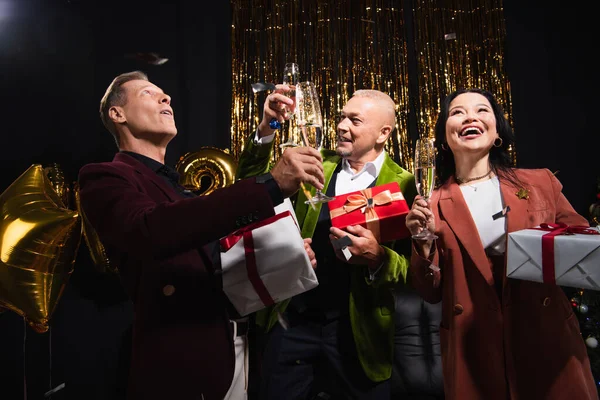 Sonrientes amigos maduros interracial celebración de champán y regalos durante la fiesta de Navidad sobre fondo negro - foto de stock