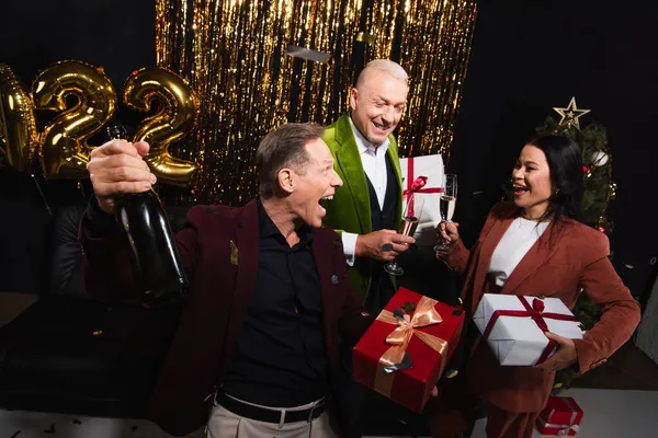 Захоплений чоловік тримає подарунок і шампанське біля міжрасових друзів з подарунками під час новорічної вечірки на чорному тлі — стокове фото