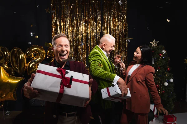 Fröhlicher Mann hält Geschenk in der Nähe von interrassischen Freunden mit Champagner während der Neujahrsparty auf schwarzem Hintergrund — Stockfoto