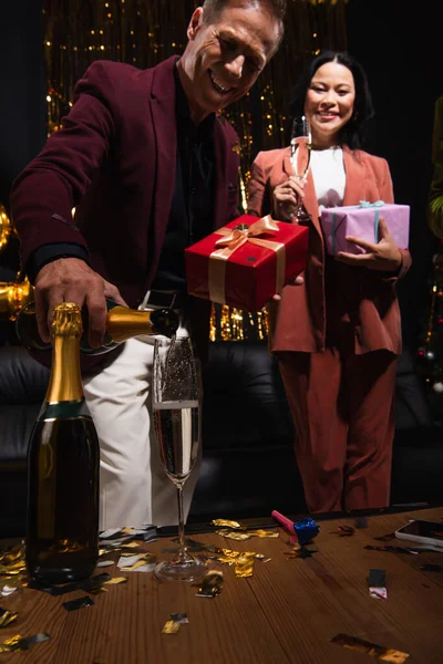 Verschwommener Mann mit Geschenk, das Champagner neben asiatischem Freund und Konfetti auf schwarzem Hintergrund einschenkt — Stockfoto