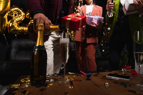 Cropped view of man pouring champagne near blurred friends with gifts and confetti during new year party on black background — Stock Photo