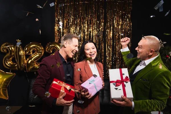 Aufgeregte Freunde mit Geschenken, die das neue Jahr in der Nähe von Konfetti und Luftballons auf schwarzem Hintergrund feiern — Stockfoto