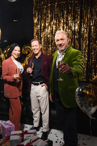 Homme mature positif tenant champagne près d'amis multiethniques, ballons et cadeaux sur fond noir — Photo de stock