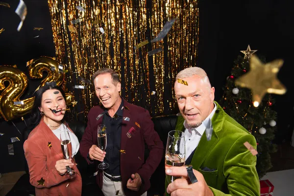 High angle view of mature interracial friends with champagne looking at camera under confetti during new year party on black background — Stock Photo