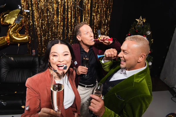 Vue grand angle de femme asiatique excitée tenant champagne près des amis avec des cornes de fête sur fond noir — Photo de stock