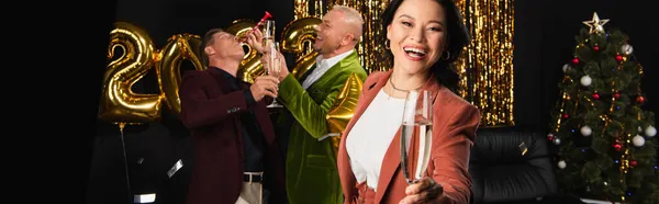 Smiling asian woman with glass of champagne looking at camera near friends celebrating new year on black background, banner — Stock Photo
