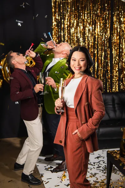Asian woman with champagne looking at camera near friends with party horn during new year party on black background — Stock Photo