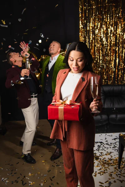 Excited asian woman holding champagne and present near friends during new year party on black background — Stock Photo