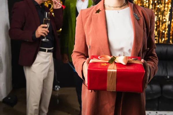 Cropped view of woman holding gift box with confetti near blurred friends on black background — Stock Photo
