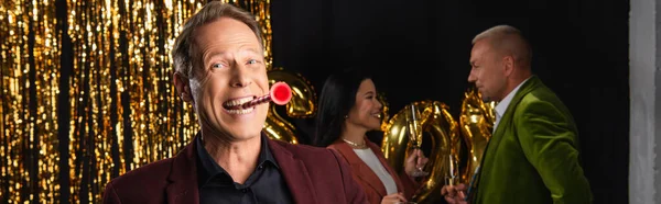 Fröhlicher, reifer Mann mit Partyhorn, der in die Kamera schaut, bei interrassischen Freunden mit Champagner und Lametta auf schwarzem Hintergrund, Banner — Stockfoto