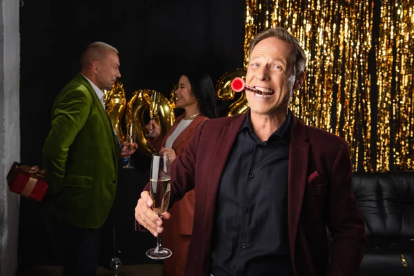 Fröhlich reifer Mann mit Partyhorn und Champagner, der in die Kamera schaut, in der Nähe von interrassischen Freunden und Lametta auf schwarzem Hintergrund — Stockfoto