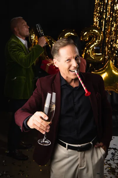 Smiling mature man with party horn and champagne looking at camera near blurred friend and festive decor on black background — Stock Photo