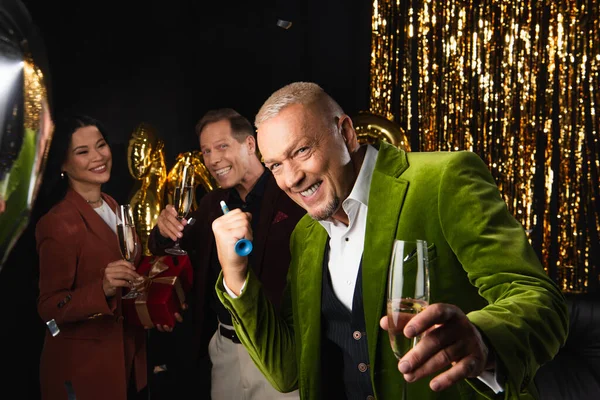 Feliz hombre maduro celebración de cuerno de fiesta y champán cerca de amigos multiétnicos y decoración festiva sobre fondo negro - foto de stock