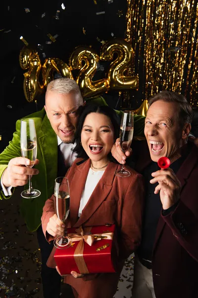 Hochwinkelaufnahme von aufgeregten interrassischen Freunden mit Champagner und Geschenk, die während der Neujahrsparty auf schwarzem Hintergrund in die Kamera schauen — Stockfoto