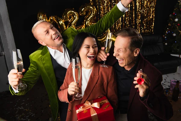 Positive multiethnic mature friends with champagne, party horn and present celebrating new year on black background — Stock Photo