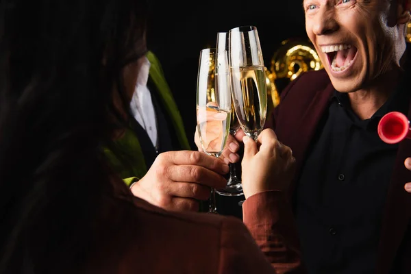 Cropped view of excited man with party horn toasting champagne with blurred friends on black background — Stock Photo