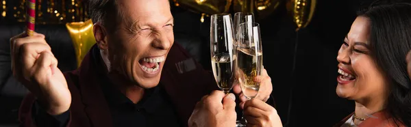Amigos multiétnicos positivos brindando con champán durante la fiesta de año nuevo sobre fondo negro, pancarta - foto de stock