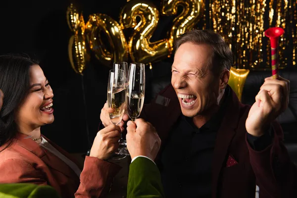 Positive man with party horn toasting champagne with multiethnic friends during new year party on black background — Stock Photo