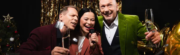 Mulher asiática cantando karaoke perto de amigos com champanhe durante a festa de ano novo no fundo preto, banner — Fotografia de Stock