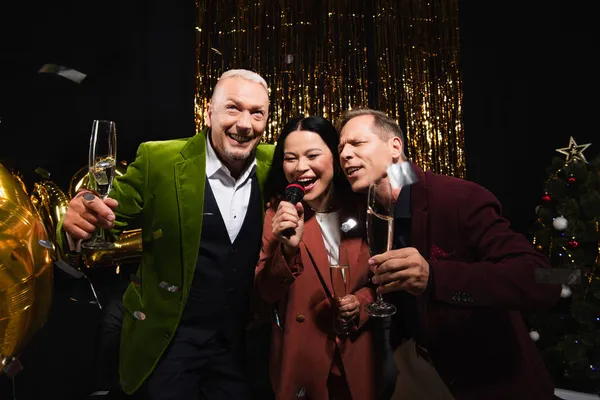 Feliz madura interracial amigos con champán cantando karaoke durante la fiesta de año nuevo sobre fondo negro - foto de stock