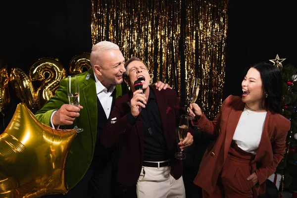 Asian woman holding champagne near friends singing karaoke during new year party on black background — Stock Photo