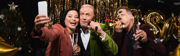 Mujer asiática tomando selfie con amigos sosteniendo champán y micrófono durante la fiesta de año nuevo sobre fondo negro, bandera - foto de stock