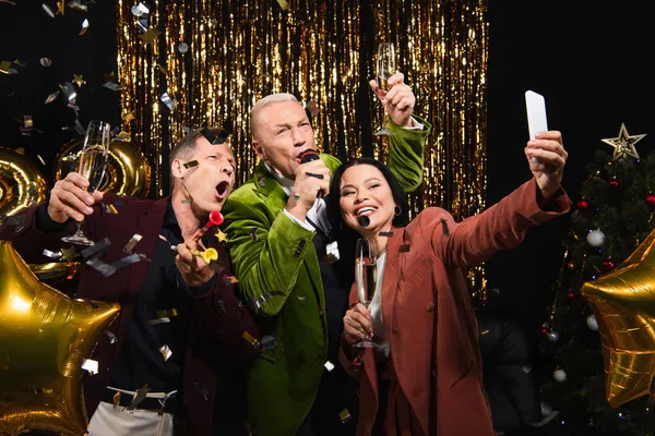 Excitados amigos inter-raciais tomando selfie e cantando karaoke durante a festa de ano novo em fundo preto — Fotografia de Stock