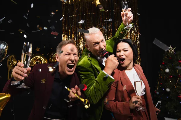 Excited multiethnic friends with champagne singing karaoke under confetti during party on black background — Stock Photo