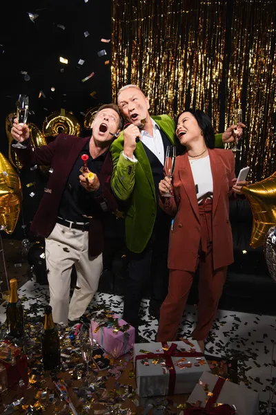 Excité amis multiethniques matures avec champagne et corne de fête chantant karaoké près décor festif sur fond noir — Photo de stock