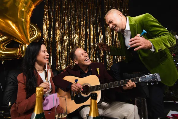 Sonriendo amigos interracial con champán tocando la guitarra acústica durante la fiesta de año nuevo sobre fondo negro - foto de stock