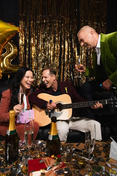 Positive interrassische Freunde mit Champagner spielen Akustikgitarre in der Nähe von Geschenken während der Neujahrsparty auf schwarzem Hintergrund — Stockfoto