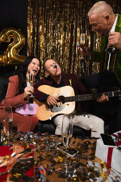 Heureux amis multiethniques avec cornes de fête et champagne jouant de la guitare acoustique lors de la fête du Nouvel An sur fond noir — Photo de stock