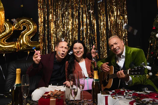 Emocionados amigos interracial con guitarra acústica celebrando año nuevo cerca de champán y regalos sobre fondo negro - foto de stock