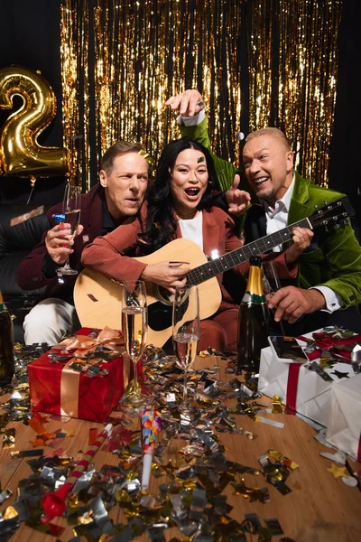 Amis multiethniques positifs jouant de la guitare acoustique près de cadeaux, champagne et confettis sur fond noir — Photo de stock