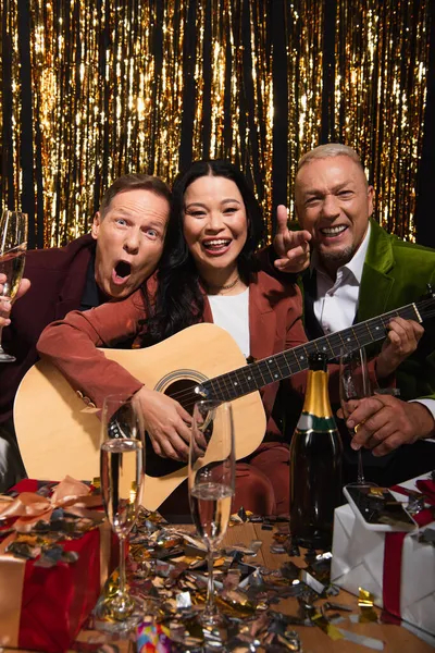 Emocionados amigos multiétnicos con champán tocando la guitarra acústica cerca de confeti durante la fiesta sobre fondo negro - foto de stock