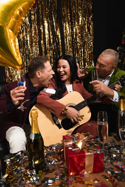 Amigos maduros interraciales con champán tocando la guitarra acústica cerca del presente y confeti sobre fondo negro - foto de stock