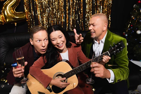 Positive interracial friends with champagne playing acoustic guitar during new year party on black background — Stock Photo