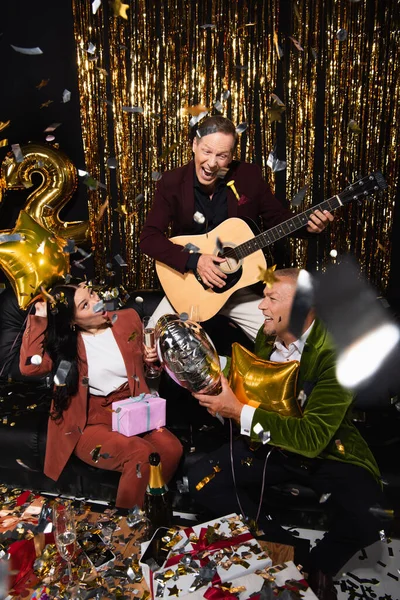 Amis mûrs multiethniques excités jouant de la guitare acoustique près du champagne et des confettis sur fond noir — Photo de stock