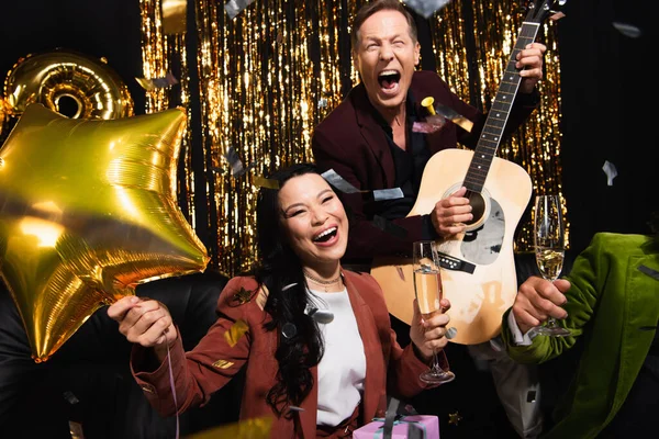 Cheerful asian woman holding balloon and champagne near friends with acoustic guitar and confetti on black background — Stock Photo