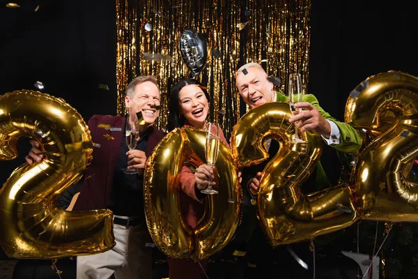 Interracial friends holding champagne near balloons in shape of 2022 numbers during party on black background — Stock Photo