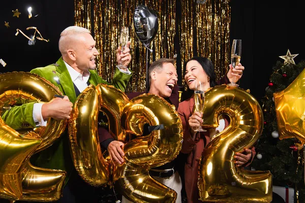 Amigos multiétnicos felizes com champanhe celebrando o ano novo perto de balões festivos em forma de 2022 números no fundo preto — Fotografia de Stock