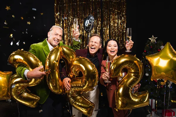 Cheerful multiethnic friends holding champagne near balloons in shape of 2022 during new year party on black background — Stock Photo