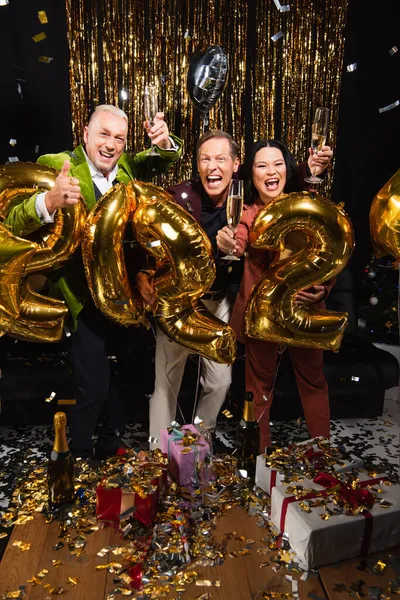 Aufgeregte multiethnische Freunde mit Champagner feiern Neujahr in der Nähe von Konfetti, Geschenken und Luftballons auf schwarzem Hintergrund — Stockfoto