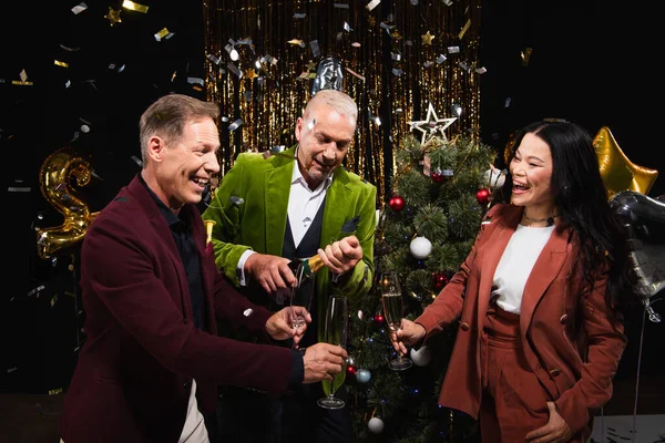 Smiling man opening champagne near interracial friends and confetti during new year party on black background — Stock Photo